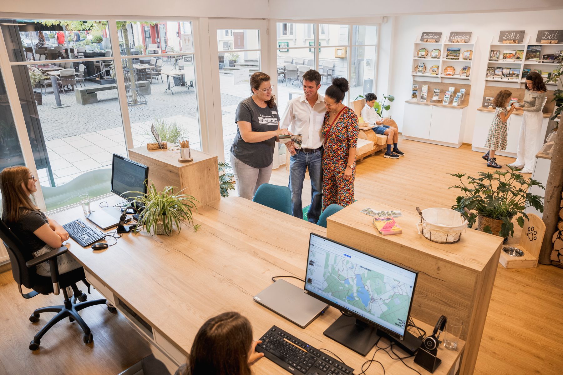 Ein modernes Büro mit großen Fenstern, die auf eine Straße hinausgehen. Mehrere Personen unterhalten sich, arbeiten an Schreibtischen oder blättern in Regalen mit Broschüren. Zwei Computer zeigen Karten an, und Pflanzen schmücken den Raum.